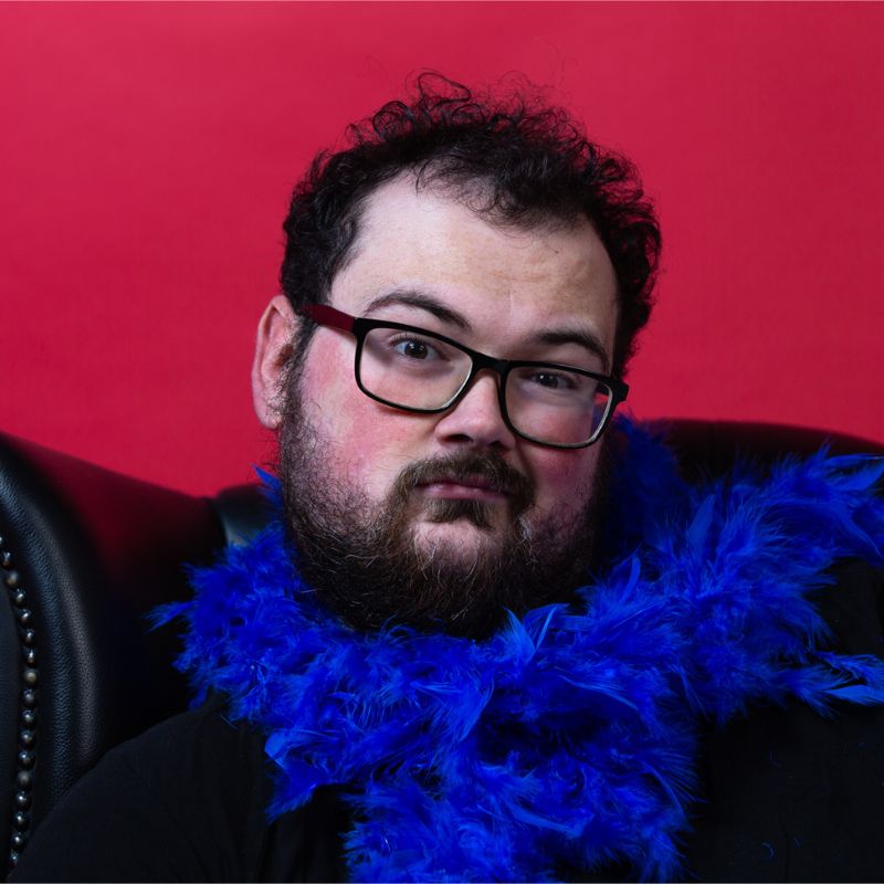 Alex Mitchell - dark haired comedian wearing black-framed glasses and blue feather boa against a red background.